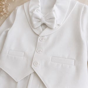 May include: A white long-sleeved jacket featuring a shawl collar, a matching textured bow tie, and three front buttons. The garment includes two horizontal welt pockets on the front, providing a structured appearance with clean lines and solid white fabric.
