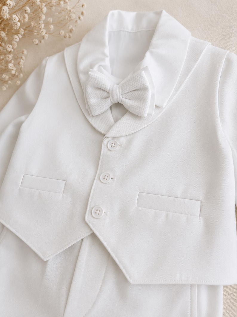 May include: A white long-sleeved jacket featuring a shawl collar, a matching textured bow tie, and three front buttons. The garment includes two horizontal welt pockets on the front, providing a structured appearance with clean lines and solid white fabric.