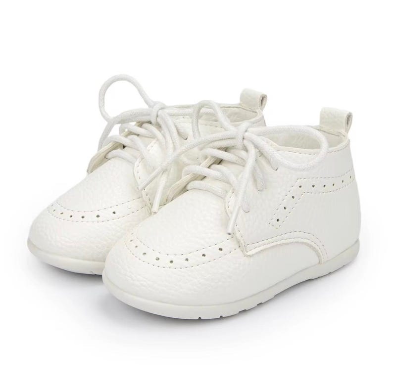 K&ouml;nnte beinhalten: Ein Paar wei&szlig;e Babyschuhe mit strukturiertem Leder-Aussehen. Die Schuhe haben ein klassisches Schn&uuml;rdesign und dekorative Perforationen. Die Schuhe sind f&uuml;r S&auml;uglinge konzipiert.