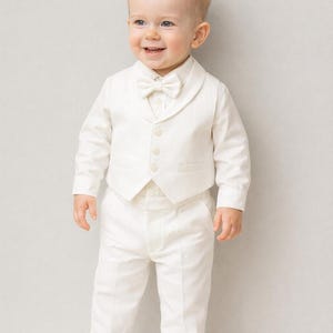 K&ouml;nnte beinhalten: Ein junges Kind in einem formellen elfenbeinfarbenen Anzug, bestehend aus Jacke, Weste, Hose und Fliege. Das Outfit wird durch wei&szlig;e Schuhe erg&auml;nzt. Das Kind l&auml;chelt, m&ouml;glicherweise f&uuml;r einen besonderen Anlass.