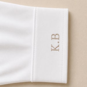 May include: A white long-sleeve shirt cuff featuring a structured band with visible stitching along the edges. The cuff is embroidered with the letters K.B in a light tan thread, positioned vertically on the fabric surface of the sleeve cuff.