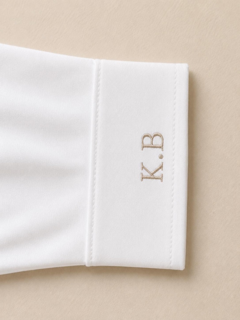 May include: A white long-sleeve shirt cuff featuring a structured band with visible stitching along the edges. The cuff is embroidered with the letters K.B in a light tan thread, positioned vertically on the fabric surface of the sleeve cuff.