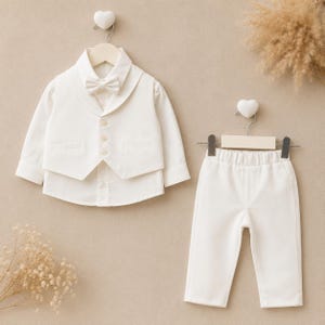 May include: A white long-sleeved button-down shirt with a matching white vest and a small bow tie, paired with white trousers. The set is displayed on two hangers, each featuring a small white heart detail at the top of the hook.