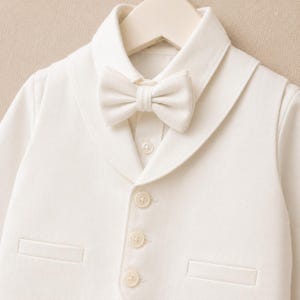 May include: An off-white vest featuring a shawl collar, a matching bow tie, and three front buttons. The garment includes two horizontal welt pockets on the lower front and is displayed on a hanger, showcasing a clean, structured design.