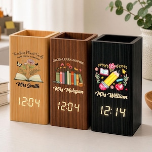 Op de afbeelding: Drie rechthoekige houten bureau-organizers met geïntegreerde digitale klokken. De lichte houten variant heeft een afbeelding van een boek en bloemen met de tekst "Teachers Plant Seeds That Grow Forever Mrs Smith". De donkerhouten variant toont boeken en de tekst "Grow-Learn-Inspire Mrs Morgan". De zwarte variant toont een potlood en appel met de tekst "Mrs William".