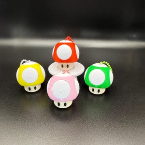 May include: Four mushroom-shaped keychains with white spots on their caps. The caps are red, yellow, pink, and green. Each keychain has a cream-colored base with two black oval eyes and a small beaded chain attached to the top of the mushroom cap.