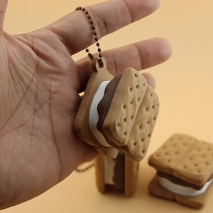 Könnte beinhalten: Ein brauner S'mores-Schlüsselanhänger mit einer Kette, mit Keks-ähnlichen Stücken, weißem Marshmallow und Schokoladenfüllung. Ein weiterer S'mores-Schlüsselanhänger und ein einzelner S'more sind ebenfalls sichtbar. Der Hintergrund ist einfarbig hellorange.
