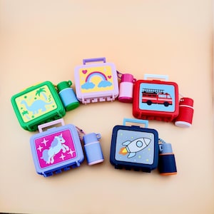 May include: Four rectangular lunch box containers with attached matching thermoses. The green box features a dinosaur, the purple box a rainbow, the red box a fire truck, the light purple box a unicorn, and the dark blue box a rocket ship.