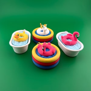 Peut inclure: Quatre porte-clés miniatures sur le thème de la piscine comprenant deux baignoires blanches avec des inserts d'eau bleu clair contenant un canard jaune et une bouée flamant rose, et deux piscines rondes avec une licorne et un flamant rose.