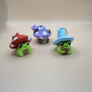 May include: Three whimsical frog figurines, each with a unique mushroom-themed design. One frog wears a red mushroom cap with white spots, another a purple mushroom house, and the third a blue wizard hat. The frogs are green with brown accents.