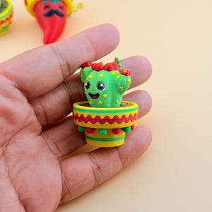 Puede incluir: Una pequeña figura de cactus verde con cara sonriente, flores rojas en la parte superior y puntos amarillos. El cactus está plantado en una maceta amarilla decorada con patrones de zigzag rojos y verdes y pequeñas esferas verdes en el borde.