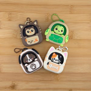 Puede incluir: Cuatro estuches para llaveros de silicona con forma de mochila, cada uno con un personaje animal diferente: un búho marrón, una tortuga verde, un cachorro marrón y blanco, y un gato blanco. Cada estuche tiene una pequeña cadena de metal.