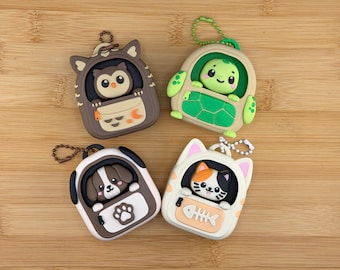 Puede incluir: Cuatro estuches para llaveros de silicona con forma de mochila, cada uno con un personaje animal diferente: un búho marrón, una tortuga verde, un cachorro marrón y blanco, y un gato blanco. Cada estuche tiene una pequeña cadena de metal.