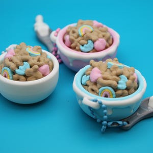 May include: Three miniature cereal bowls filled with brown cereal, pink hearts, blue stars, and rainbow marshmallows. The bowls are white, pink, and blue, with spoons and beaded handles. The background is a solid blue.