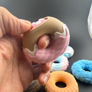 Puede incluir: Una colección de juguetes antiestrés con forma de dónut y superficies texturizadas. Un juguete es de color tostado con glaseado rosa y detalles blancos, mientras que otros son azules, naranjas o degradados, todos con un patrón acolchado.