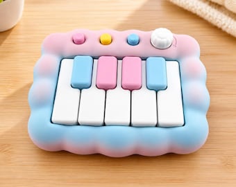 Puede incluir: Un piano de juguete rectangular con un marco suave y redondeado en degradado de rosa a azul. Cuenta con cinco teclas blancas, dos teclas azules, dos teclas rosas, tres pequeños botones circulares en rosa, amarillo y azul, y un dial de color blanco.