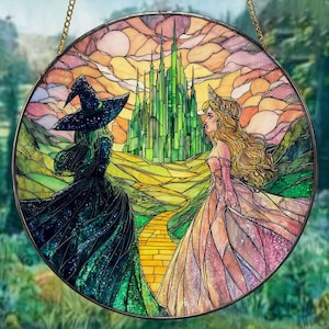 May include: Circular stained glass panel depicting a scene from The Wizard of Oz. The artwork shows the Wicked Witch of the West and Glinda facing the Emerald City. The panel has a gold chain for hanging and is framed in a dark metal.