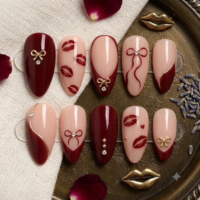 May include: A set of ten almond-shaped press-on nails featuring a color palette of deep burgundy and nude. The designs include hand-painted red lips, delicate gold-toned bows, thin gold lines, and small embedded rhinestones on various nails.