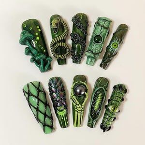 May include: A collection of ten long, green press-on nails with various designs. The designs include mushrooms, snakes, and other fantasy-themed elements. The nails are a glossy green with black accents, creating a unique and artistic look.