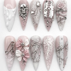May include: A set of ten long, pointed artificial nails in various designs. The nails feature a pale pink and white color scheme with embellishments like a skull, lace, chains, and floral accents. Some nails have a glossy finish, while others have a matte texture.