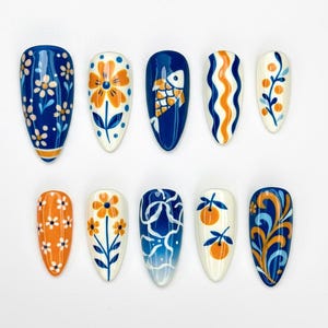 May include: A set of ten almond-shaped false nails with various hand-painted designs. The designs include flowers, fish, and abstract patterns in shades of blue, orange, and white. The nails are arranged on a white surface.