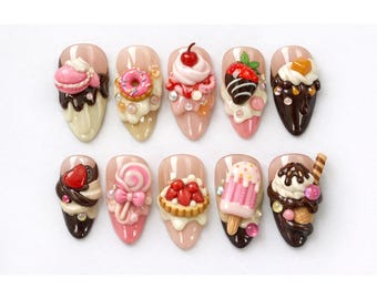 May include: A collection of ten artificial nails, each adorned with miniature dessert-themed decorations. Designs include a macaron, donut, cherry, chocolate-covered strawberry, and ice cream cone, all in vibrant colors.
