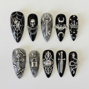 May include: A set of ten long, pointed false nails with a gothic theme. Designs include a spiderweb, skull, key, bat, crown, cross, padlock, ornate jewel, dagger, and snake, all in silver against black and gray backgrounds.