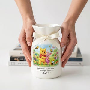 May include: A white ceramic vase with a ruffled rim and a cream-colored ribbon tied around the neck. The front features a colorful illustration of Winnie the Pooh and Piglet, bees, and flowers, with the text: Sometimes the smallest things take up the most room in your heart.