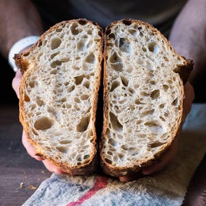 May include: Two halves of a loaf of bread, revealing the interior texture. The bread has a golden-brown crust and a light, airy interior with large holes. The bread is held by a person.