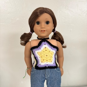 May include: A doll with brown hair styled in pigtails, wearing a crochet halter top featuring a yellow star pattern with white, lavender, and black borders, paired with blue denim jeans. The doll has brown eyes and a light skin tone.