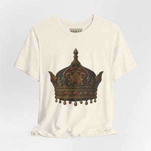 Könnte beinhalten: Cremefarbenes T-Shirt mit einer detaillierten Illustration einer königlichen Krone. Die Krone ist mit roten, grünen und goldenen Juwelen verziert und hat eine dunkelrote und grüne Oberseite. Auf dem Shirt steht das Wort "MIRAI".
