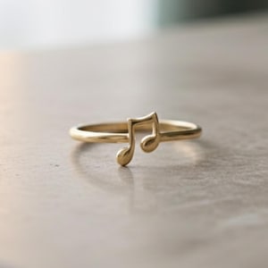 May include: A delicate gold ring featuring a musical note design. The ring's band is thin and smooth, with the musical note symbol prominently displayed. The ring is set against a neutral background, highlighting its elegant simplicity.