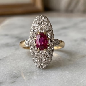 Può includere: Un anello vintage con un rubino ovale centrale circondato da un alone di piccoli diamanti scintillanti. L'anello ha una fascia in oro con accenti di diamanti su entrambi i lati del rubino e del gruppo di diamanti. L'anello è posto su una superficie di marmo.