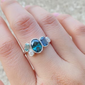 May include: A silver ring featuring an oval, teal-colored gemstone as the centerpiece, flanked by smaller, rectangular and round gemstones in shades of teal and blue. The ring is set against a neutral background.