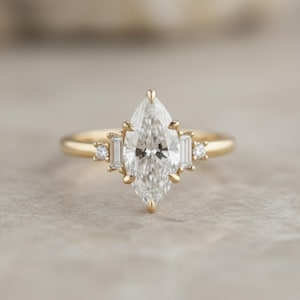 Peut inclure: Une bague de fiançailles en or avec un diamant central de taille marquise. Des diamants rectangulaires plus petits ornent les côtés de la pierre principale, sur un anneau fin.
