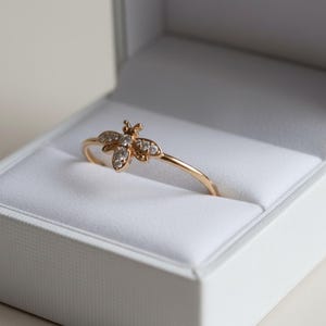May include: A gold ring in a white jewellery box. The ring has a bee design with small, clear gemstones. The ring is delicate and sits on a white, padded surface inside the box. The box is open, revealing the ring.