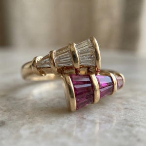 May include: A gold bypass ring featuring two tapered sections of channel-set baguette-cut gemstones. One section contains clear white stones, while the other section contains deep pink-red stones, both set within a polished yellow gold band.