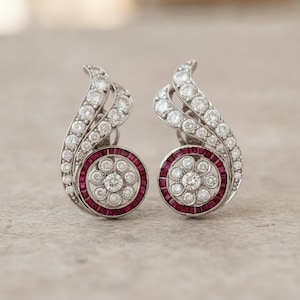 May include: A pair of silver-toned earrings featuring a swirling design set with numerous small round white diamonds. Each earring includes a central circular motif with a flower-like arrangement of diamonds surrounded by a ring of deep red rubies.