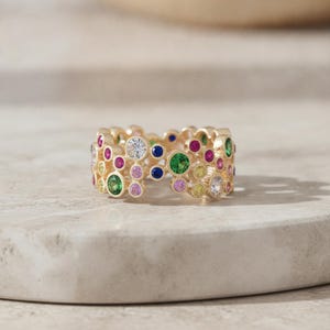 May include: A gold band ring featuring a cluster of bezel-set, multi-coloured round gemstones in various sizes. The stones include shades of green, pink, blue, yellow, and clear white, arranged in an organic, bubble-like pattern across the surface.