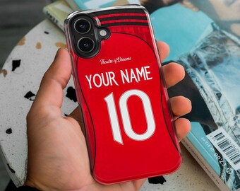 May include: A red phone case featuring a jersey design with the text "YOUR NAME" and the number "10" in white. The case also has the words "Theatre of Dreams" in a script font. The case is clear with a black and red trim.