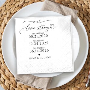 May include: A white square napkin with the text "our love story" in elegant script. The napkin lists three dates: 05.21.2020, 12.24.2025, and 06.14.2026, along with the names Emma & Hudson. The napkin is placed on a white plate.