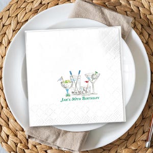 May include: White square paper napkin with a diamond pattern, featuring illustrations of cocktails and golf clubs. The text "Jax's 30th Birthday" is printed in green. The napkin is placed on a white plate with a woven placemat.