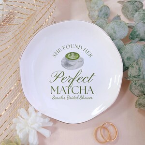 Op de afbeelding: Witte keramische plaat met een gouden rand, met een matcha latte illustratie en de tekst "She Found Her Perfect Matcha" en "Sarah's Bridal Shower" in het groen. Twee gouden ringen staan op de voorgrond.