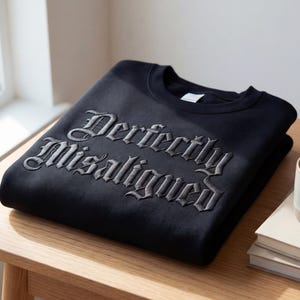 Op de afbeelding: Een zwarte sweatshirt met de geborduurde tekst "Perfectly Misaligned" in een gotisch lettertype. Het gevouwen kledingstuk ligt op een houten tafel, naast een kopje koffie en een stapel boeken.