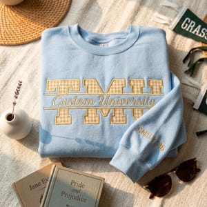 Pode incluir: Camisola azul clara com texto bordado "Custom University". As letras têm um padrão xadrez vichy bege. A manga mostra o nome "Emily 26" bordado a ouro. Livros e óculos de sol rodeiam a camisola.