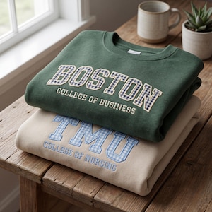 Pode incluir: Duas camisolas empilhadas numa superfície de madeira. A camisola superior é verde escuro com "BOSTON COLLEGE OF BUSINESS" bordado em xadrez vichy branco e azul. A camisola inferior é bege com "TMU COLLEGE OF NURSING" bordado em xadrez vichy azul claro.