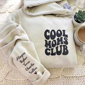 Könnte beinhalten: Ein cremefarbener Kapuzenpullover mit dem Text "COOL MOMS CLUB" in Schwarz auf der Vorderseite. Der linke Ärmel zeigt Namen in schwarzer Schrift. Eine kleine Sukkulente in einem weißen Topf und eine Tasse Kaffee sind im Hintergrund. Der Hoodie liegt auf einer cremefarbenen Strickdecke.