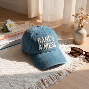 Puede incluir: Gorra de béisbol azul con el texto bordado "CARL'S A MESS" en blanco. La gorra está sobre una alfombra blanca con flecos. Una margarita, libros y gafas de sol están en el fondo.