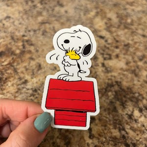 Snoopy and Woodstock Hugging on Red Doghouse – Peanuts Vinyl Sticker for Laptops, Water Bottles, Journals – 3"x1.75" Waterproof Decal