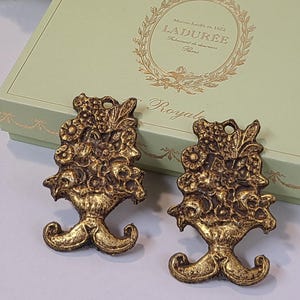 Peut inclure: Deux pièces décoratives en métal doré ornées. Chaque pièce présente un motif floral avec une base en forme de vase et une boucle en haut. Les pièces sont posées sur une boîte vert clair avec des lettres dorées.